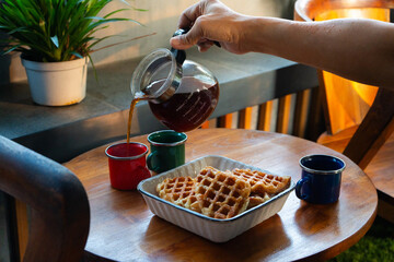 A person pours hot coffee from a glass pot into a cup on a cozy wooden table. The scene features...