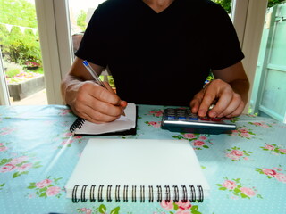 Man sitting at table using a calculator and notepad in a living room with garden in the background.