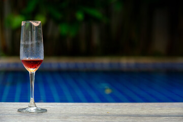 a wine glass with lipstick mark and some red wine water left at swimming pool side  and blurred green plants in background