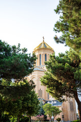 Obraz premium A scenic view of the Holy Trinity Cathedral of Tbilisi, also known as Sameba Cathedral, partially hidden among lush green trees. The golden dome and stone façade shine through the natural frame