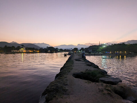 Scenic Waterfront Dusk with Tranquil Reflections, Illuminated Buildings, Mountain View, and People Relaxing on Rustic Stone Pier 