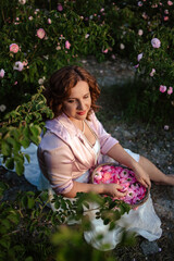 Romantic woman in light dress with curly hair sitting in blooming rose garden with picnic basket, enjoying summer nature outdoors, floral field lifestyle, relaxation and beauty in countryside scenery
