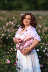 Romantic woman in light dress with curly hair sitting in blooming rose garden with picnic basket, enjoying summer nature outdoors, floral field lifestyle, relaxation and beauty in countryside scenery