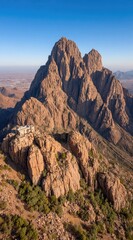 An aerial view of a rugged, rocky mountain illuminated by warm sunlight, with a small structure nestled on a plateau and sparse vegetation
