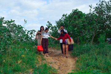 Four teenage girls descend a dirt slope surrounded by trees and grasses, holding hands to support...