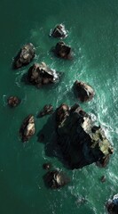 Overhead view of dark rocks surrounded by turquoise sea water. Waves crash gently against the stones, creating a serene, isolated scene