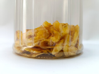 A clear jar containing sweet banana chips with a yellow lid isolated on a white background.