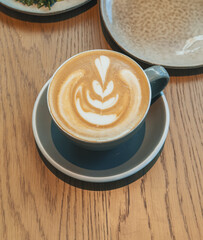 A cup of cappuccino with a decorative latte art design is served on a saucer on a wood table.