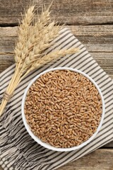 Wheat grains in bowl and spikes on wooden table, flat lay