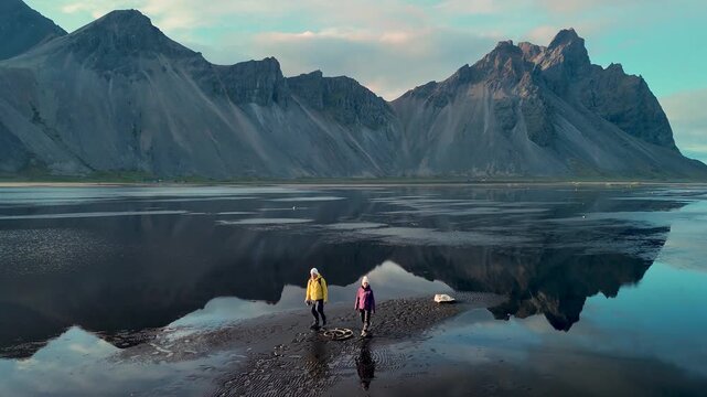 Two adventurers clad in bright outerwear stand on a serene black sand beach in Iceland, surrounded by majestic mountains and calm waters,S tokksnes Cape, and Vestrahorn Mountain