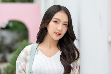 Asian Young woman with long dark hair wearing white top and pastel cardigan posing outdoors with soft smile and natural makeup, surrounded by blurred greenery and pink background