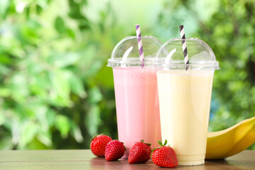 Tasty milkshakes in plastic cups and ingredients on wooden table outdoors, space for text