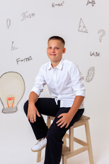 Thoughtful schoolboy in uniform sitting on wooden stool, student portrait with science and math formulas on white background, education and learning concept
