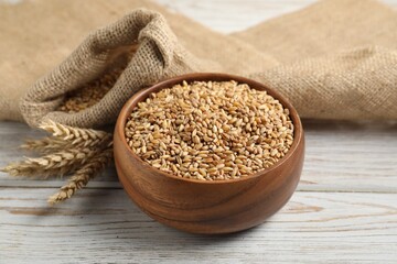 Wheat grains in bowl, spikes and burlap sack on light wooden table, closeup