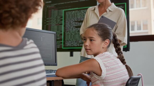 Cute diligent schoolgirl with pigtail sitting by desk in front of computer with codes on screen and looking at classmate during discussion of rules of decoding information at lesson
