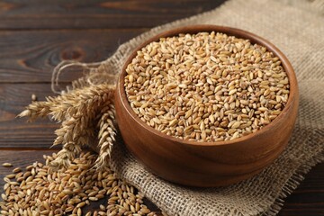 Wheat grains in bowl, spikes and burlap cloth on wooden table, closeup