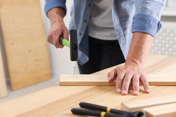 Man hammering nail into wooden plank indoors, closeup. Space for text