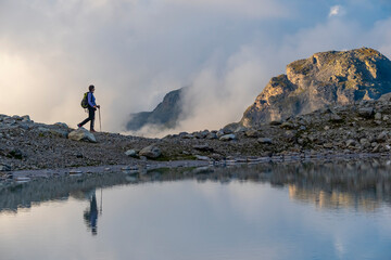 A person stands on rocky terrain, gazing at a stunning turquoise mountain lake surrounded. Dressed...