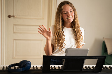 Front view of happy young woman taking online piano lessons at home using tablet waving her hand,...