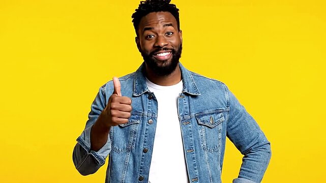 An AfricanAmerican man on a yellow background gives a thumbsup He is wearing a denim jacket over a white Tshirt