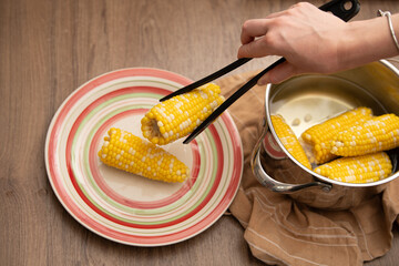 A hand uses tongs to transfer steaming corn on the cob from a pot to a vibrant plate. The scene...