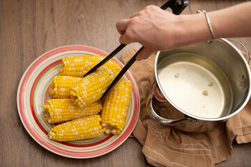 A person uses tongs to serve boiled corn on the cob from a colorful plate in a warm kitchen. Fresh ingredients enhance the cooking experience for a comforting meal