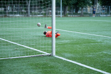 A young goalkeeper in bright orange gear dives towards the ground to save a soccer ball as players engage in an afternoon match on a well-maintained field surrounded by trees