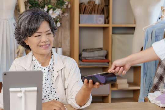 Senior asian tailor processing cashless payment with mobile phone from adult asian female client inside boutique studio modern digital transaction retail checkout small business service
