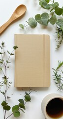 Beige notebook surrounded by greenery and a wooden spoon, coffee cup, on a white background