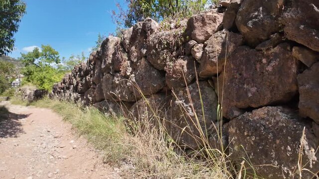 A cinematic slow-motion 4K shot captured with DJI Osmo Pocket 3 along a mountain trail. A stone wall lines the right side of the path, while trees grow on the left, with sky visible above. Perfect for