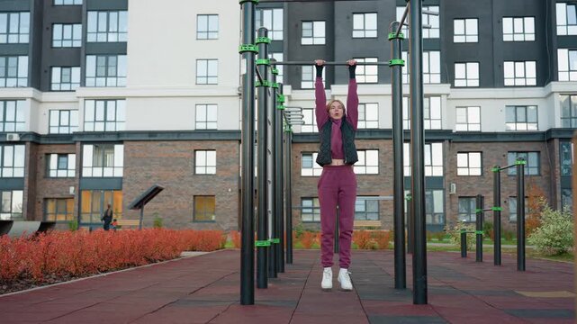 Outdoor urban park workout scene featuring female athlete in hoodie and vest hanging from pull up bar and lifting legs to engage core muscles and build strength, endurance and balance.