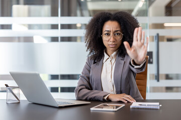 Obraz premium Portrait of an African American woman sitting at a desk in an office and showing a stop and warning sign to the camera with her hand
