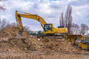Yellow crawler excavator moving rubble and soil at demolition site, with trees and construction equipment in the background.