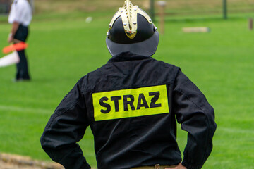 Firefighter wearing black uniform and helmet with visible STRAŻ label standing on a field during public event. © Adam