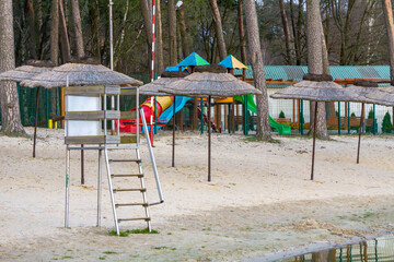 Sandy beach by lake with lifeguard tower, thatched umbrellas and colorful playground slides for children in recreation area