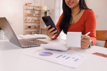 Asian woman manages finances at home, using a calculator on phone and laptop while reviewing...