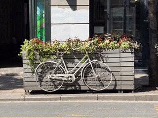 A vintage white bicycle is fixed to a wooden planter with flowers on a city street. Urban decoration, lifestyle, and creativity express charm, culture, and outdoor cafe atmosphere.