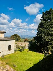 Tranquil courtyard with stone walls, lush greenery, and distant rolling hills under a bright blue sky dotted with clouds.
