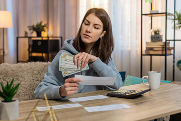 Young Caucasian woman sitting on sofa couch counting money dollar while reviewing bills. Adult girl...