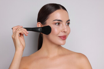 Beautiful woman applying makeup with brush on light grey background