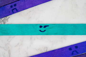 Aqua-colored popsicle stick with a winking smiley face drawn on it, lying diagonally across a marble surface and surrounded by purple sticks featuring sad faces.