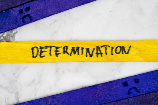 Yellow stick with handwritten word Determination placed between two purple sticks with sad face drawings on white marble background.