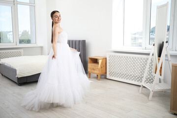 Dreams come true. Woman wearing beautiful wedding dress looking at herself in mirror indoors
