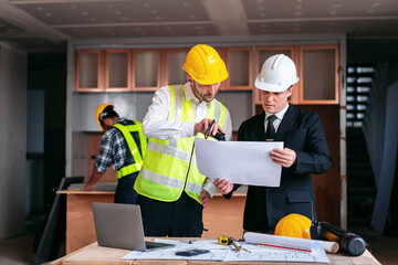 Three men are standing around a table with blueprints and a laptop