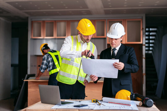 Three men are standing around a table with blueprints and a laptop
