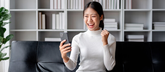 Excited Young Asian Woman Celebrating Success Looking at Smartphone on Sofa At Home Happy Win Online Bet Mobile App Message Good News Achievement