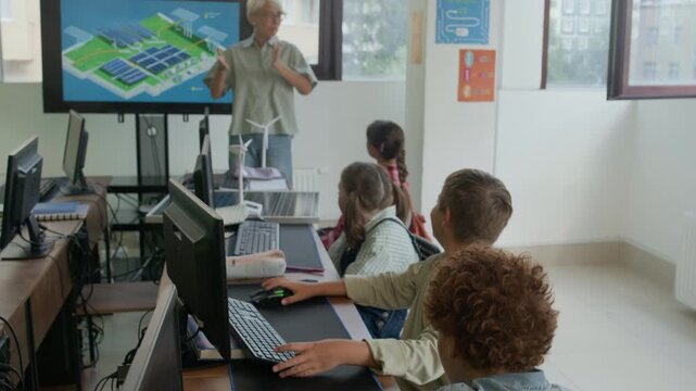 Young teacher standing by interactive board with solar panels and explaining group of diligent schoolkids how and where can be used alternative sources of energy