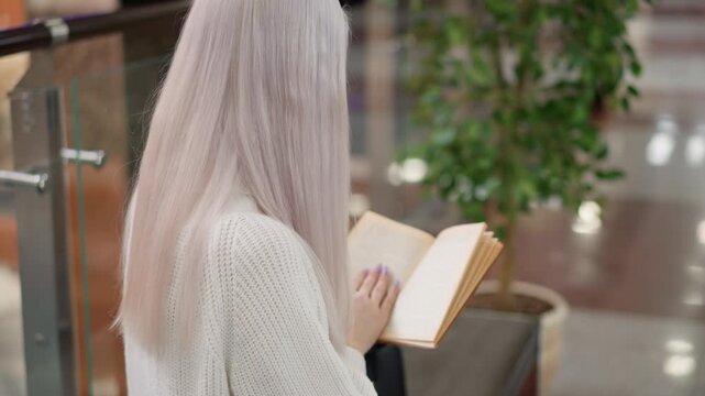 back view of youthful adult seated on bench in sweater turning pages of brown bound novel intently reading in upscale mall near potted tree during quiet reading moment with blur modern interior