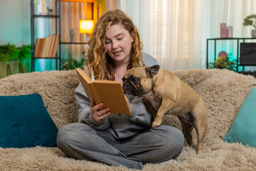 Young relaxed Caucasian redhead woman reads book aloud to pug dog on home sofa with soft voice and...