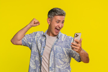 Middle-aged smiling man smartphone with joy excitement clenching fist in victorious celebration after winning online game or receiving prize notification. Young happy guy isolated on yellow background